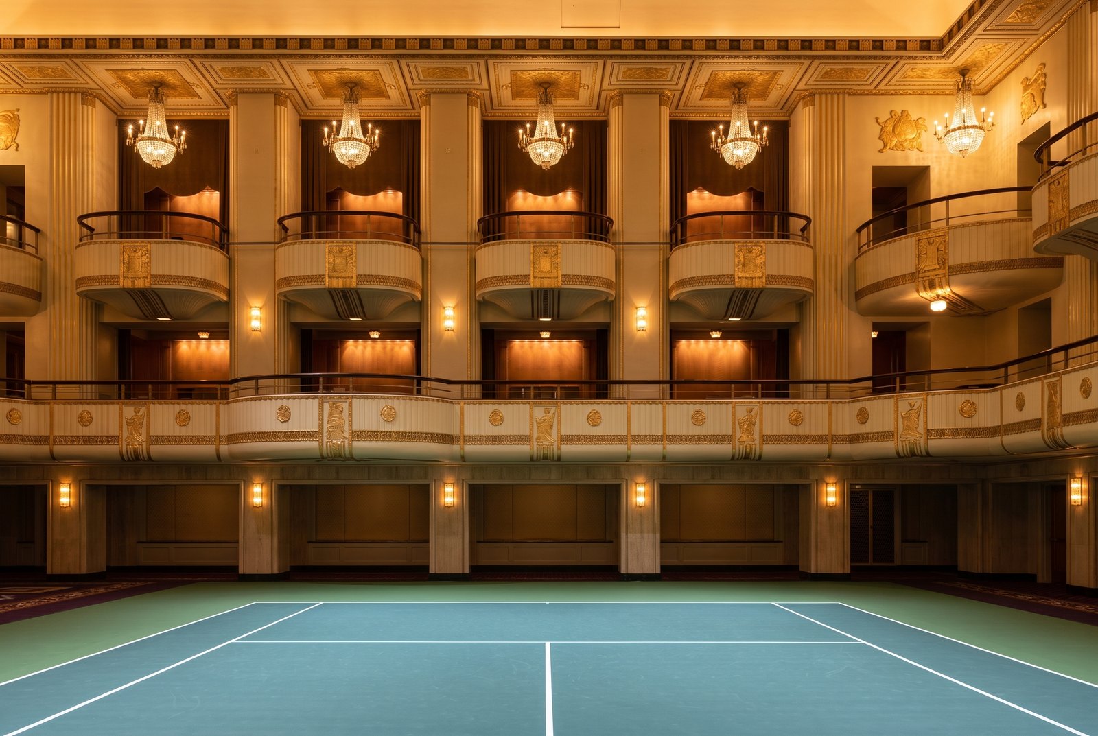 Two tiers of curved Art Deco balcony boxes overlooking a tennis court laid on a ballroom floor, chandeliers hanging between them